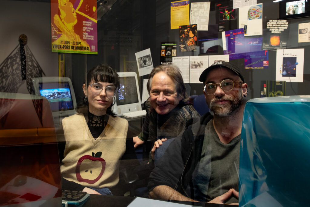 Trois personnes sont assises derrière une vitre, entourées d'anciens ordinateurs et d'affiches rétro sur le numérique. À gauche, une femme avec des lunettes. Au centre, un homme sourit. À droite, un homme avec une casquette et des lunettes regarde vers l'objectif. La scène est éclairée par des reflets sur la vitre, ajoutant une ambiance immersive.